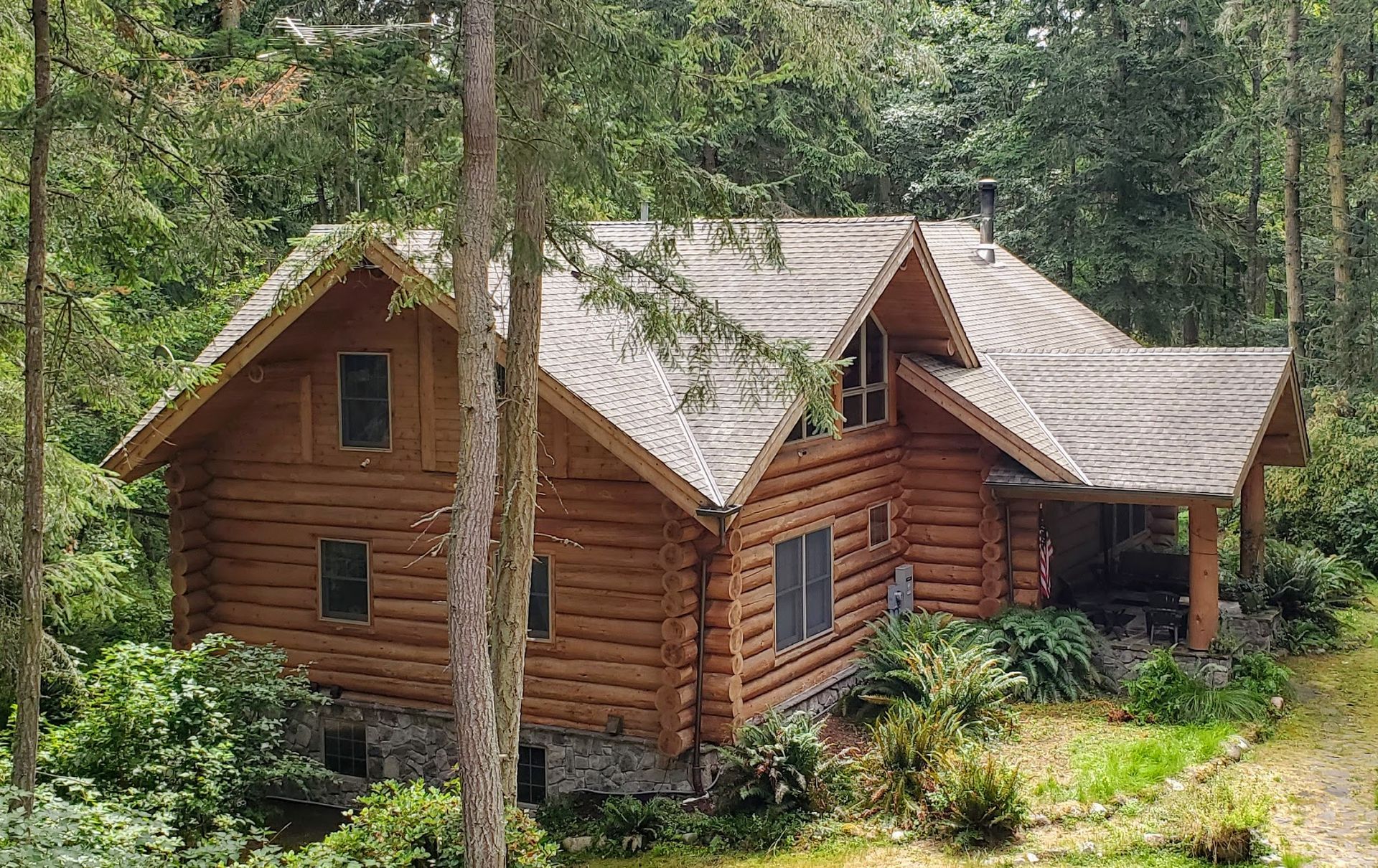 Log cabin nestled in a forest, featuring wood walls, clean, moss-free gabled roof, and porch with plants.