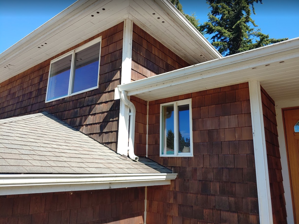 Brown shingle-clad house with white trim, gutters, and windows on a sunny day.
