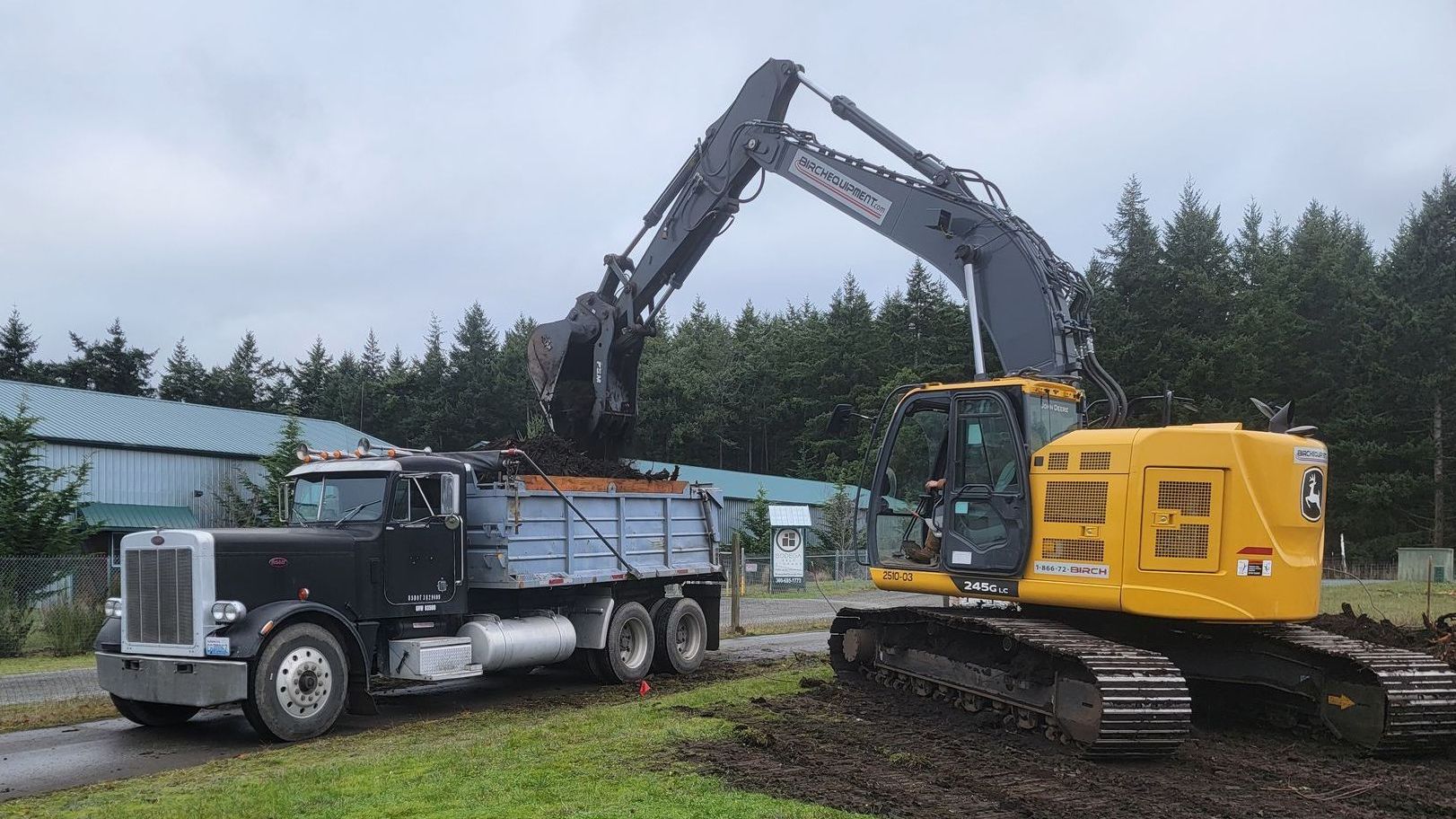 Excavator loading a black dump truck with dirt on a cloudy day; green trees and building in the background.