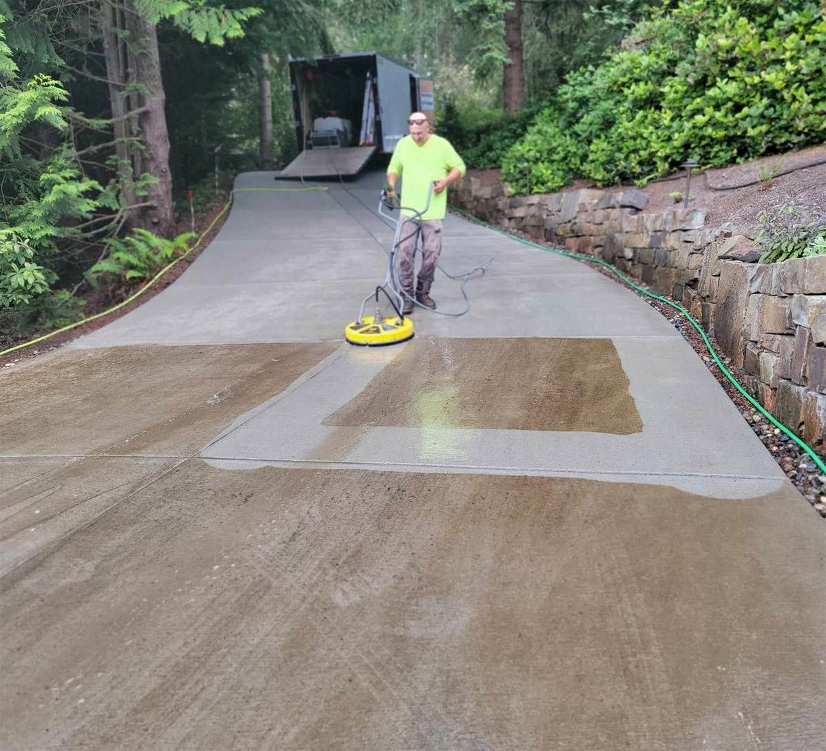 Man power washing a concrete driveway with a rotary surface cleaner; trailer in the background.