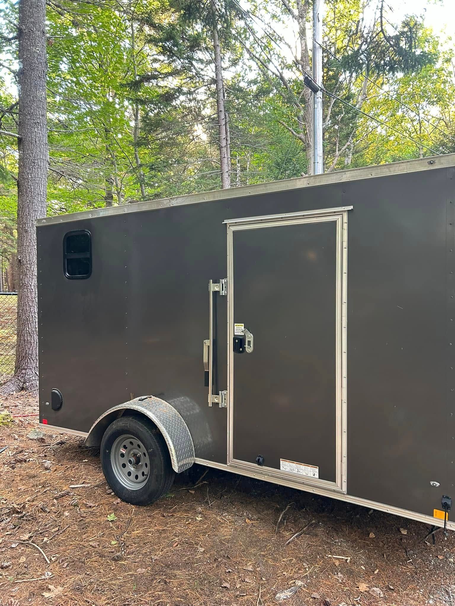 Brown cargo trailer with a door and wheel on a forest floor.