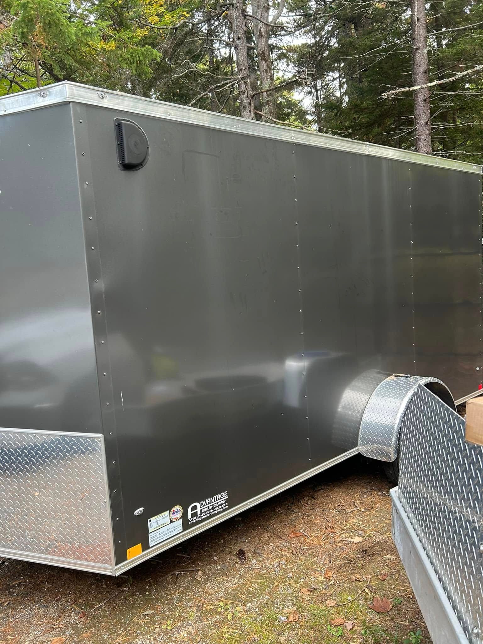 Gray enclosed cargo trailer with a diamond-plate front, parked outdoors near trees.
