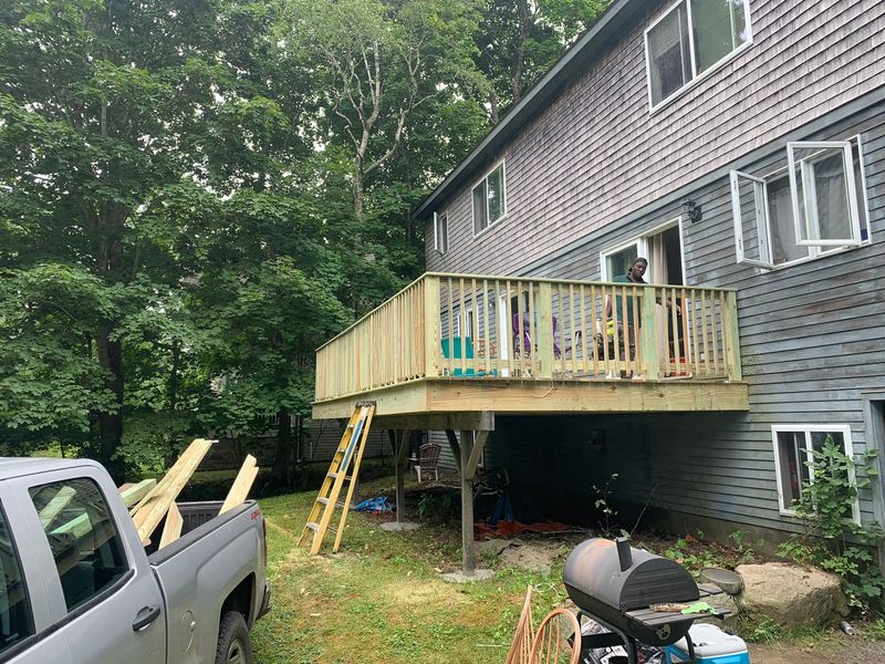 Newly built wooden deck on a two-story house, gray siding. Construction materials and truck in foreground.