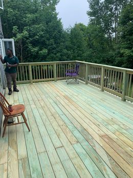 A person stands on a new wooden deck with a railing, surrounded by trees and two chairs.