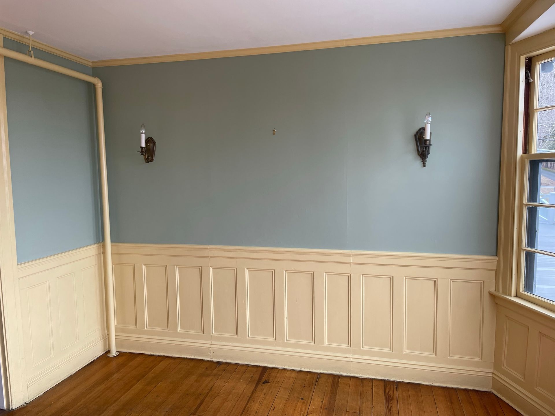 Room with blue-green walls, tan wainscoting, two sconces, and a window.