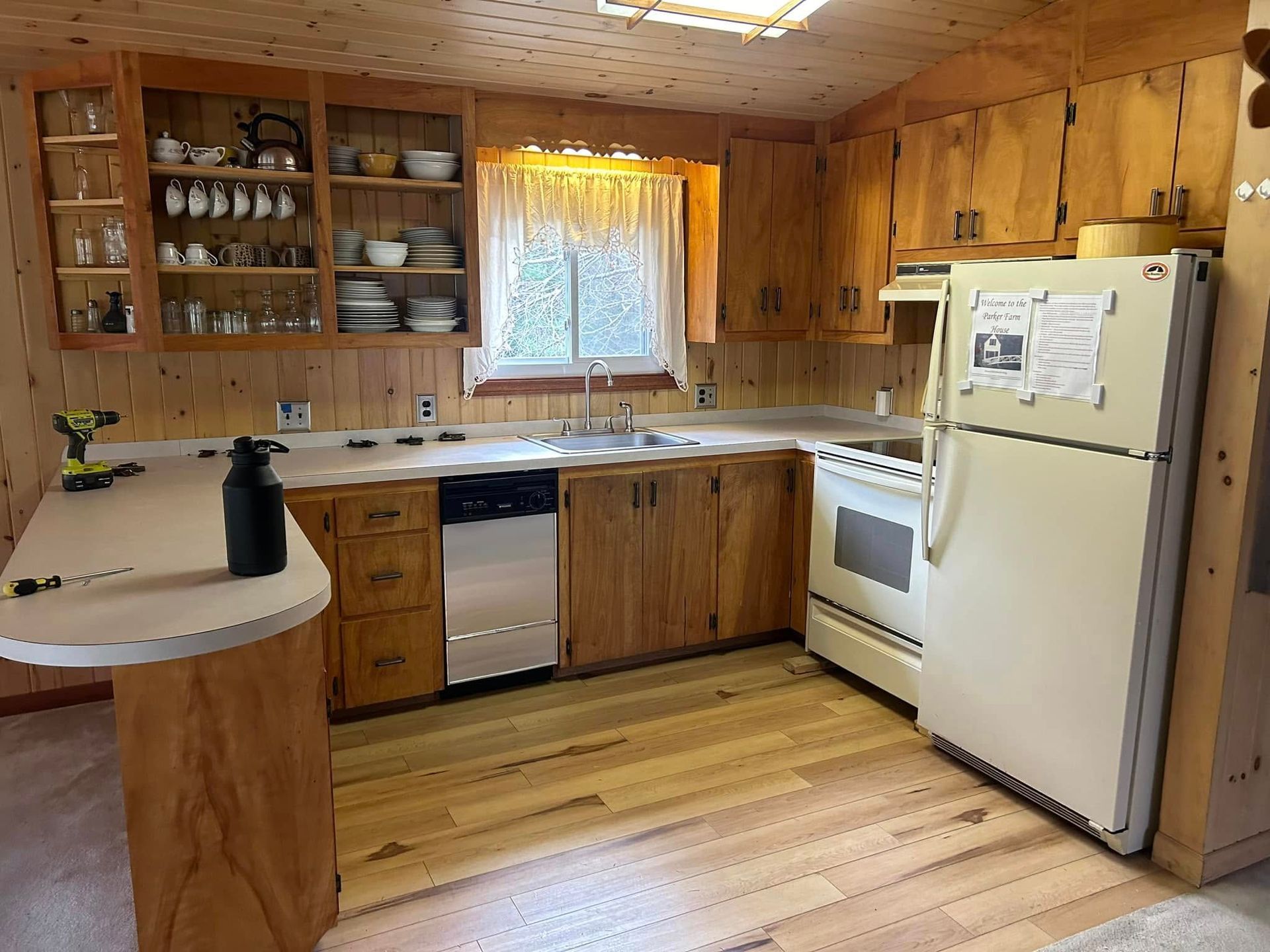 A rustic kitchen with light wood cabinets, a white stove and refrigerator. A small window is over the sink.