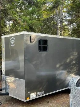 Gray enclosed trailer with a window and logo, parked outdoors with trees in the background.
