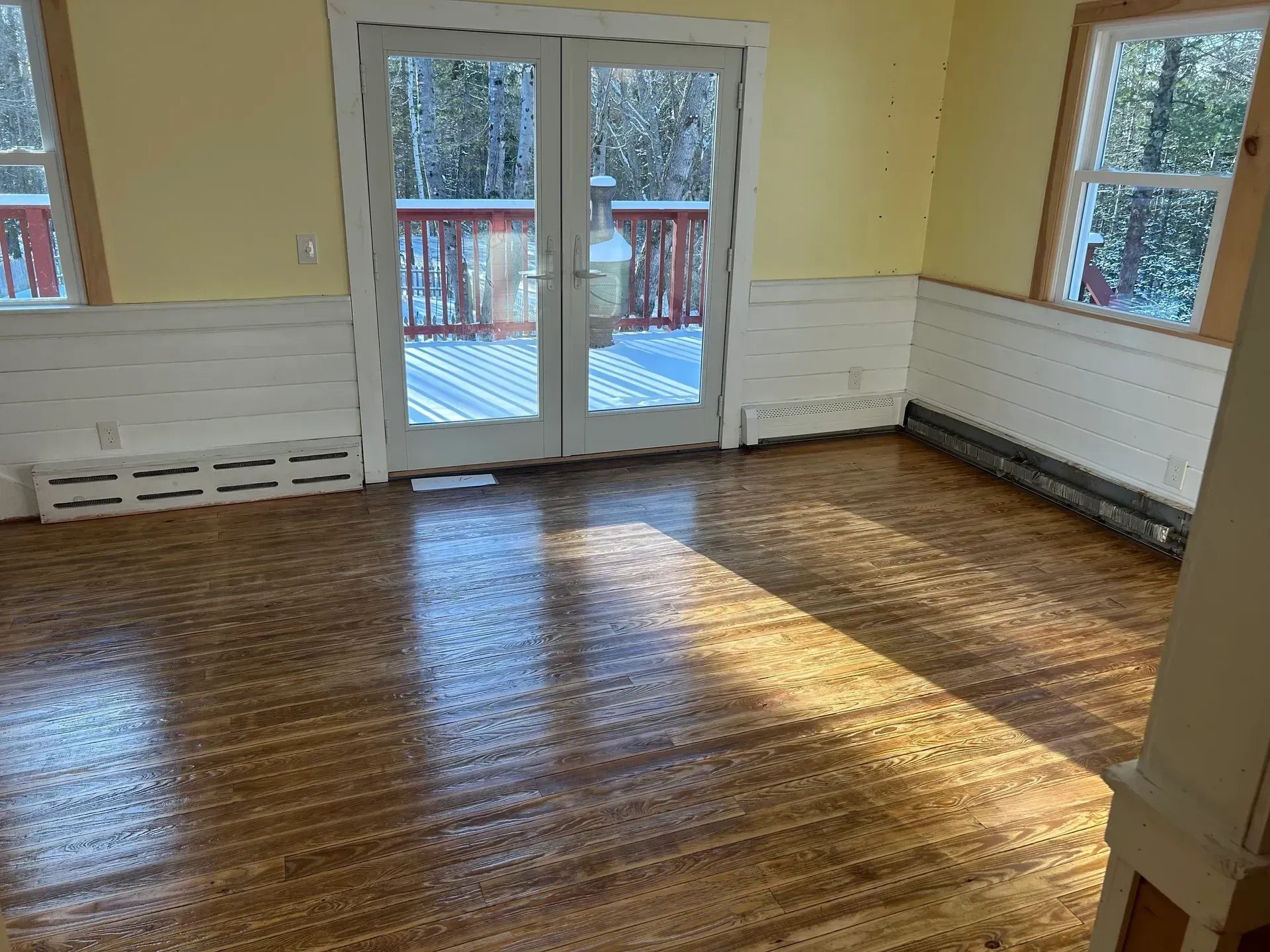 Room with wood floor, yellow walls, white wainscoting, and snow-covered deck visible through glass doors.