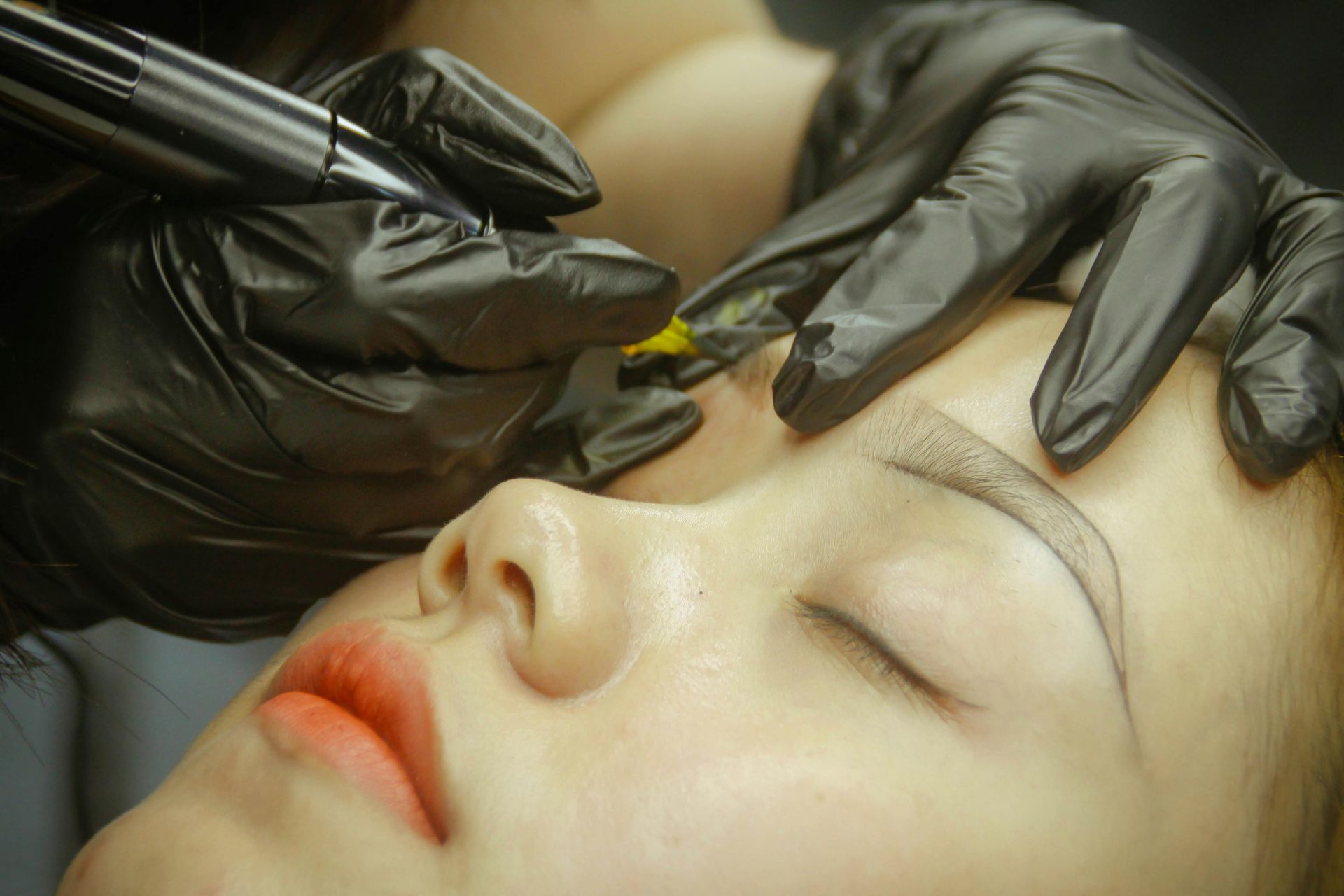 Person receiving eyebrow microblading with gloved hands using a tool.
