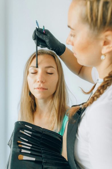 Makeup artist applying makeup to a client's eyebrow. Both have their eyes closed.