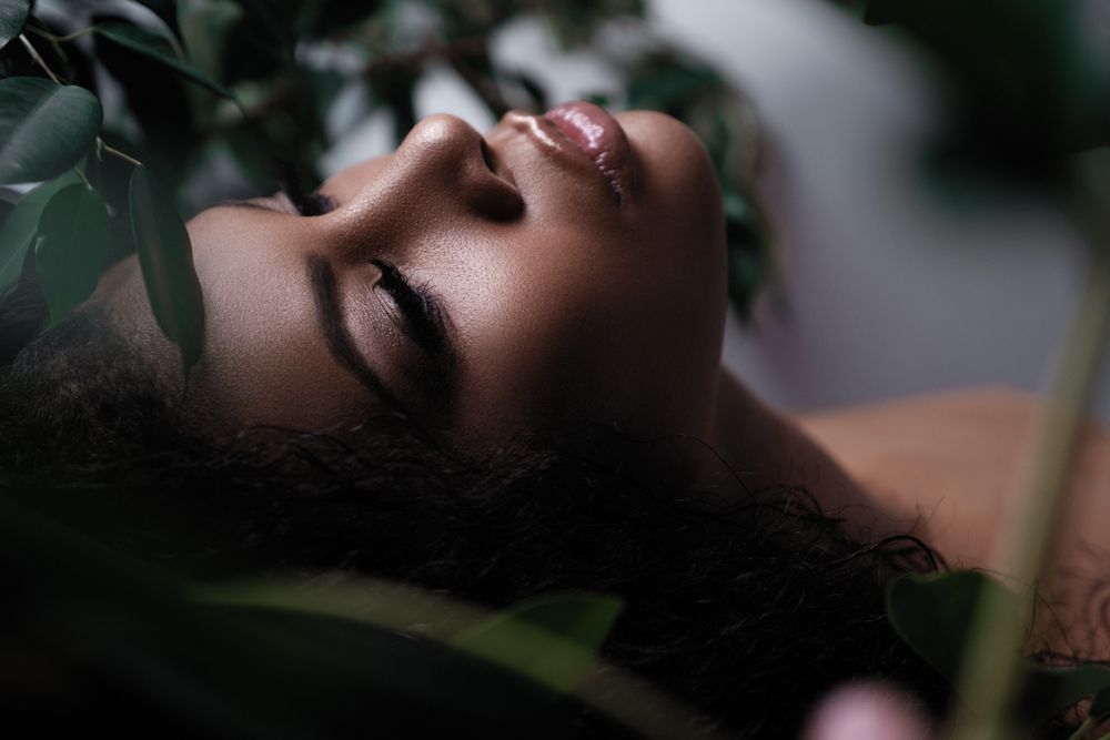 Woman's face with closed eyes, surrounded by greenery. Soft lighting on skin.