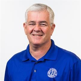Man in blue polo shirt smiles, wearing glasses. White background.