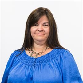 Woman with dark hair in a blue top and a necklace, smiling at the camera against a white background.