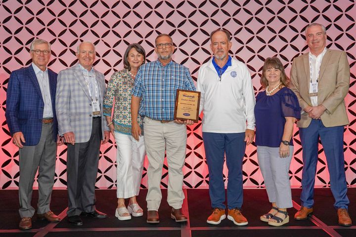 Two men in blue shirts pose in front of a banner with a logo.