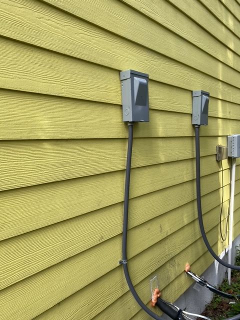 Two gray electrical boxes mounted on yellow siding, with black cables running downward.