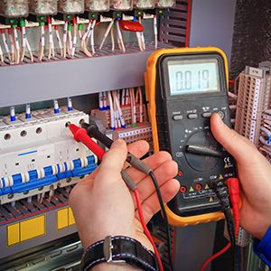 Electrician using a multimeter to test wiring in a control panel; the meter shows a reading.