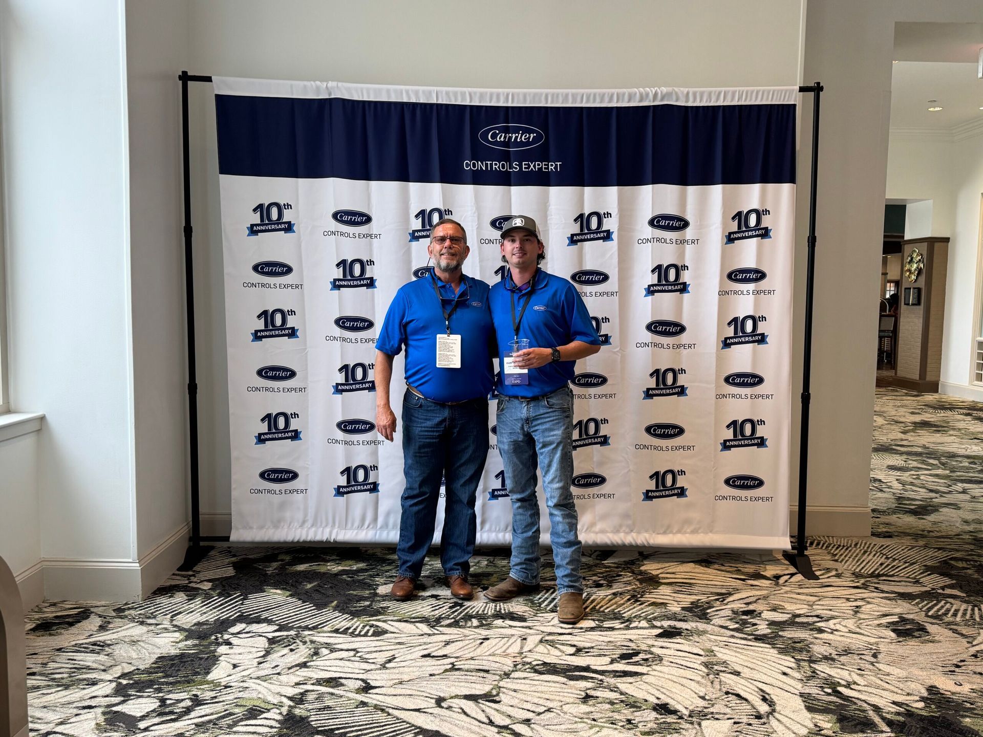Two men in blue shirts pose in front of a banner with a logo.