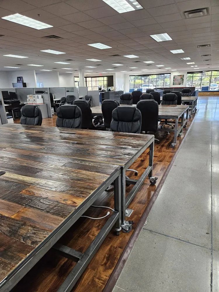 A modern office space featuring long rustic wooden desks with black ergonomic chairs on a polished wood floor.