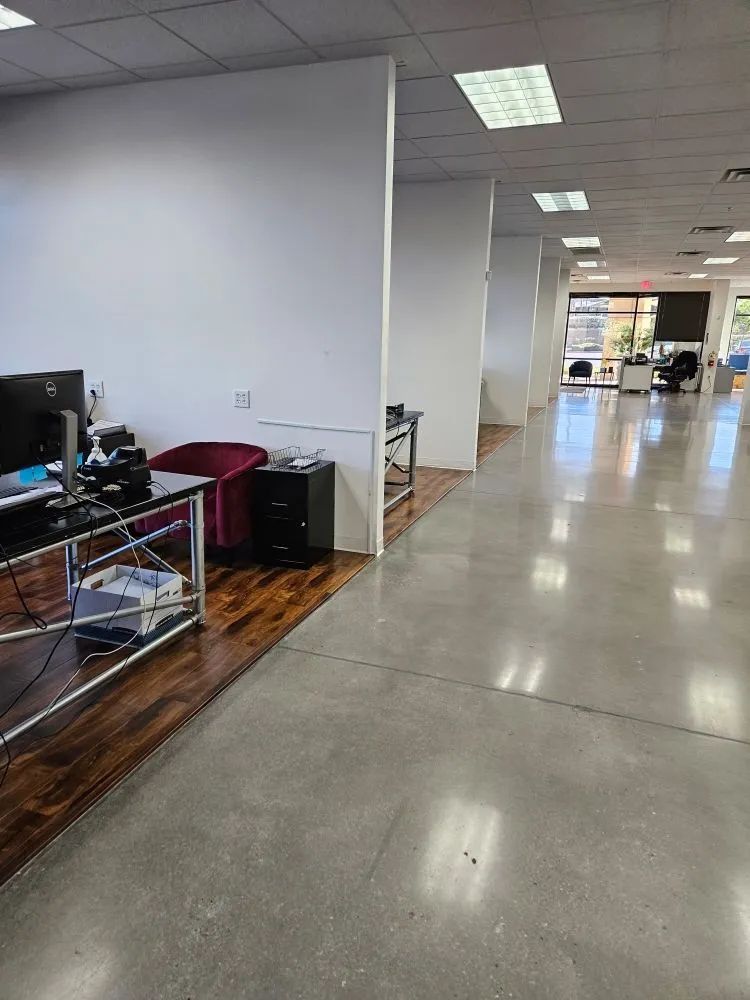 An office hallway with white partitions and a polished concrete floor, featuring a desk area with wood-patterned flooring.