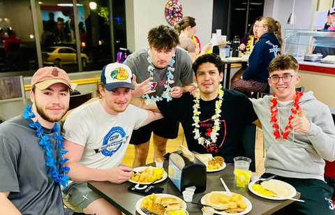Five young people with leis sit at a table eating, smiling and posing for a photo.