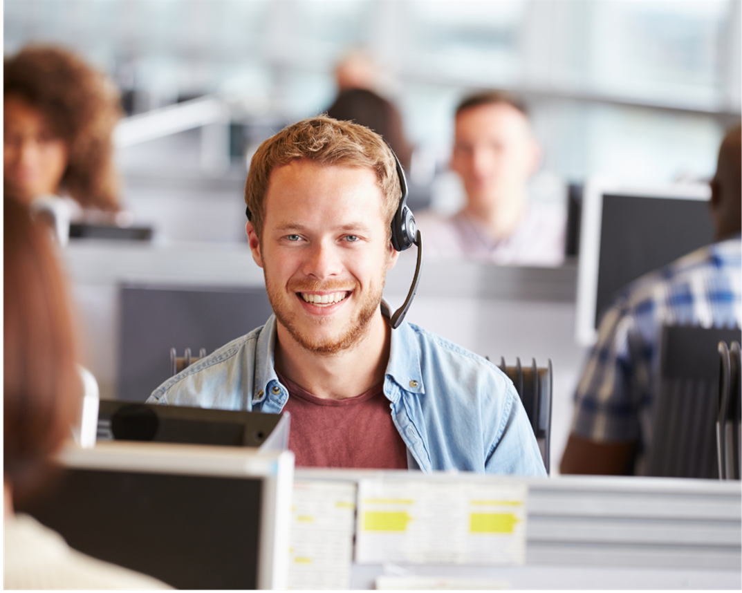 Man with headset smiles, working at a desk in a busy office, people blurred in background.