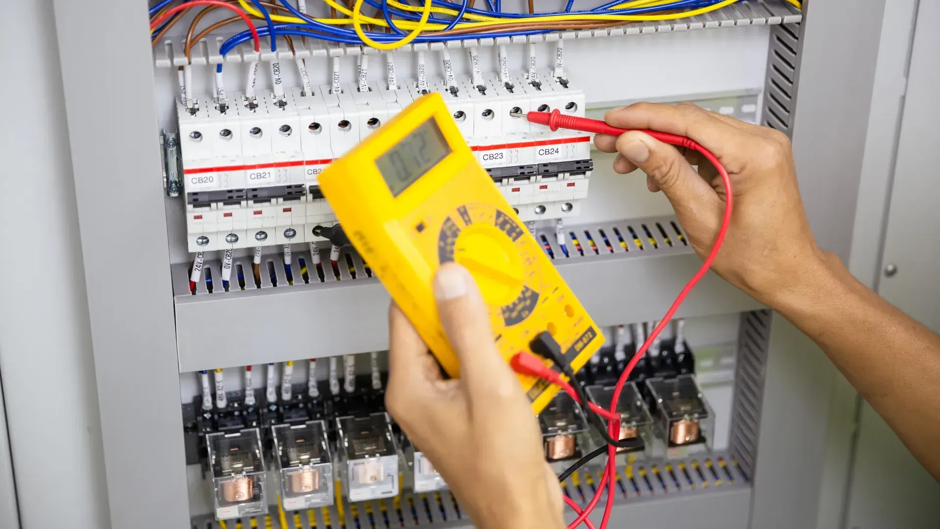 Person using a yellow multimeter to test electrical wiring inside a gray control panel.