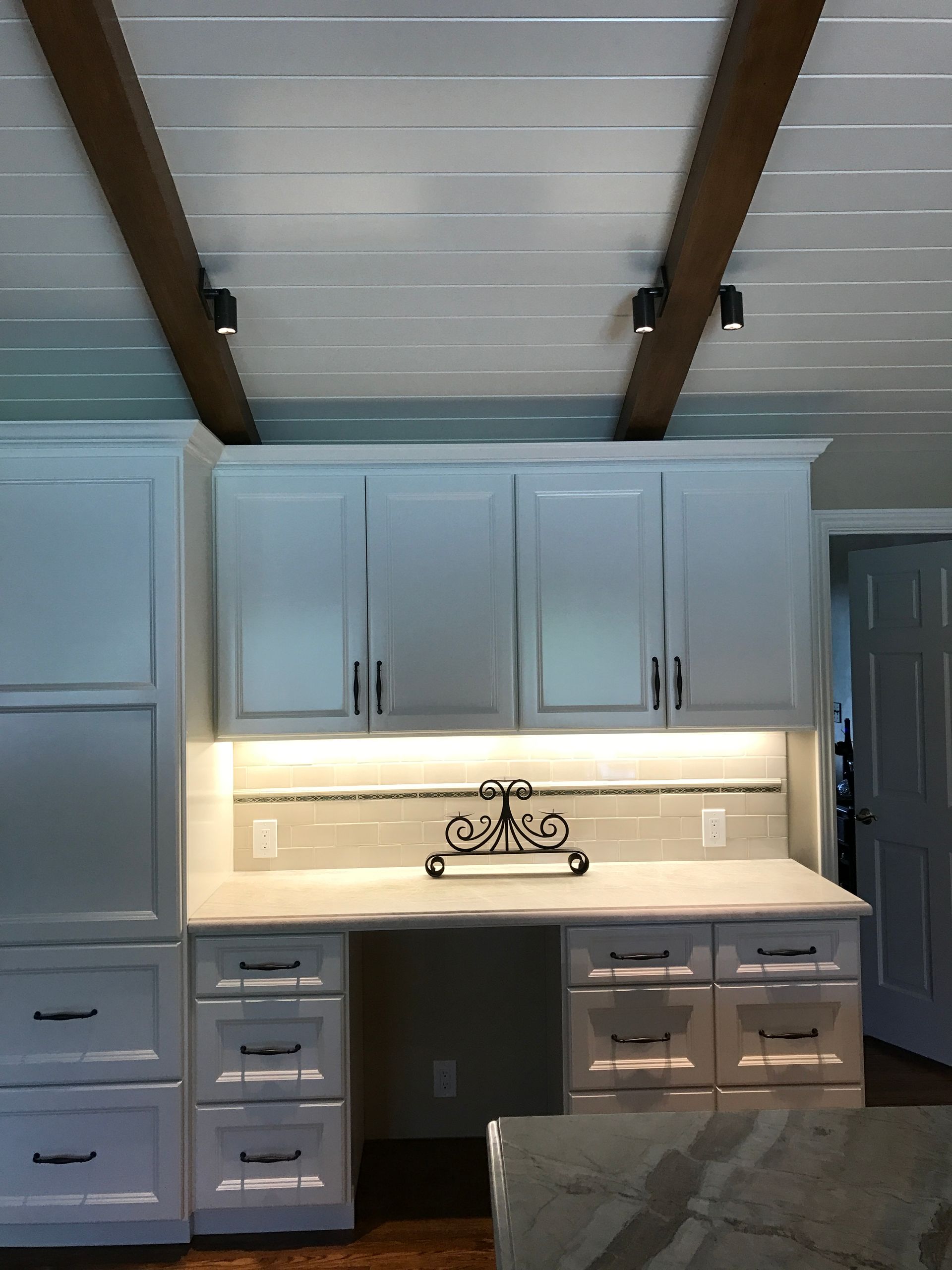 White cabinets with a built-in desk against a white-beamed ceiling. Track lighting and black hardware are visible.
