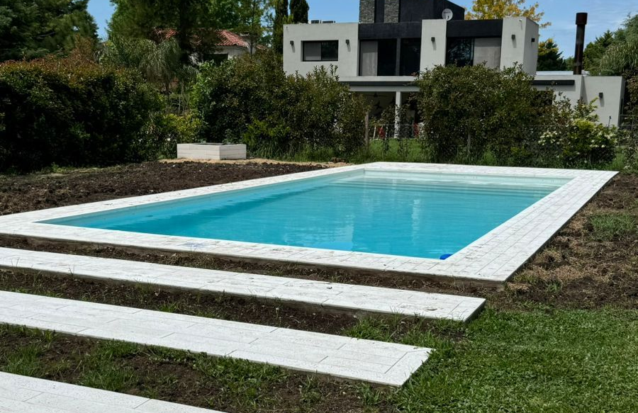 Piscina rectangular con borde de azulejos blancos; escaleras que bajan desde el césped. Casa moderna al fondo.