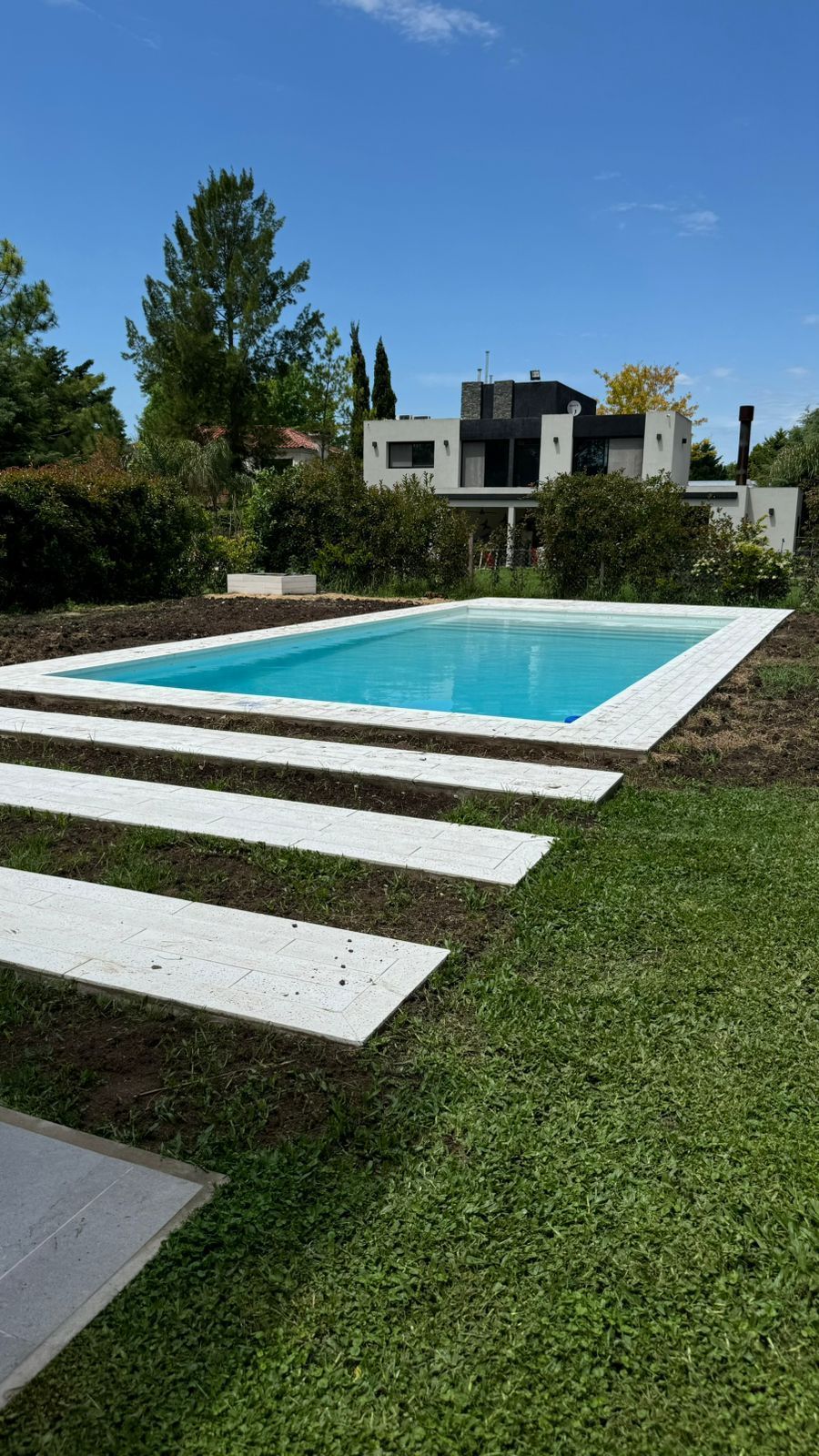 Piscina rectangular con escalones de piedra blanca, césped y casa moderna bajo un cielo azul.