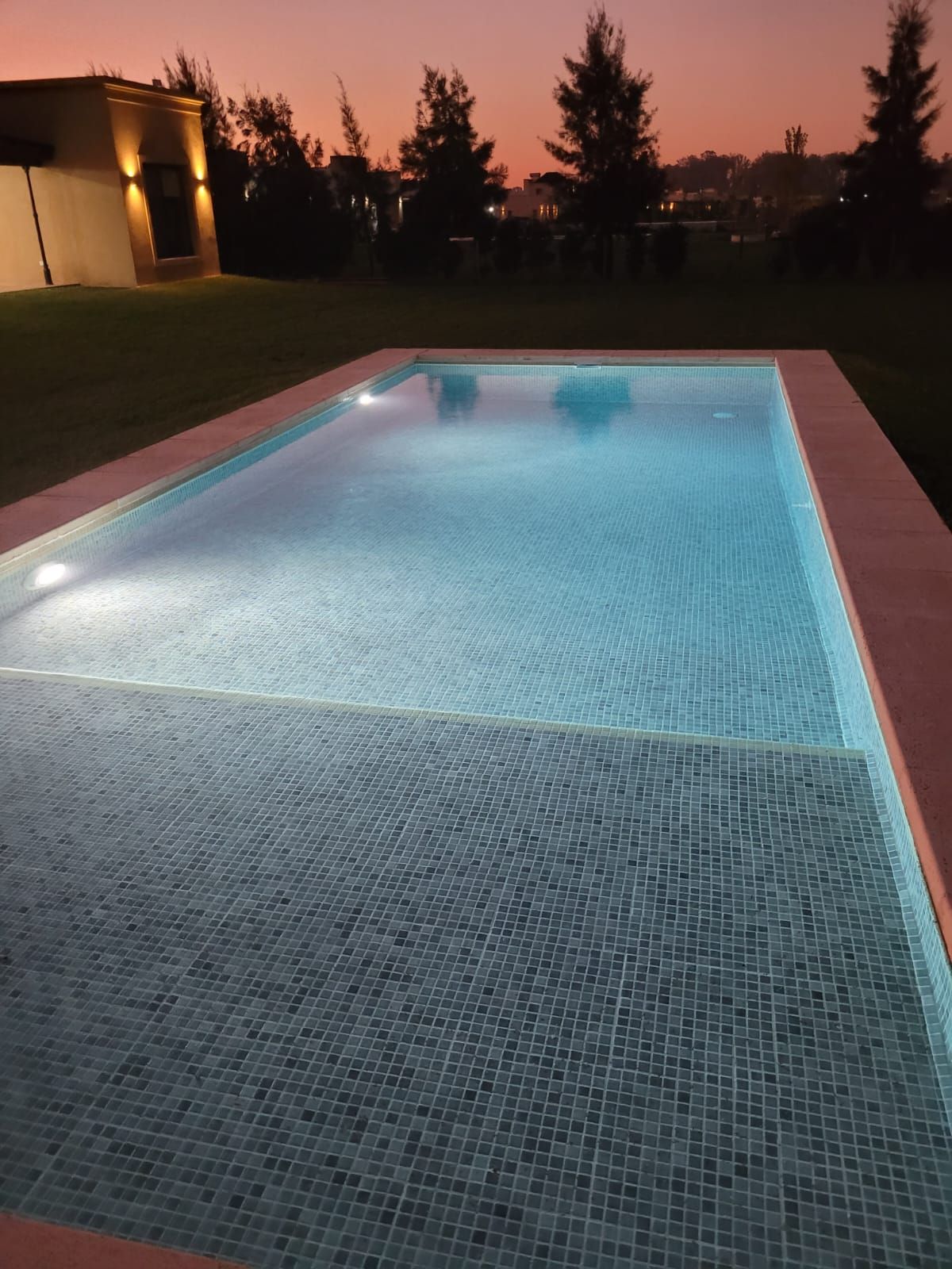 Piscina rectangular al anochecer, con escalones incorporados. Cielo rosa suave y agua iluminada.