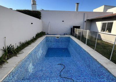 Piscina vacía rectangular de azulejos azules en un patio trasero, rodeada de paredes blancas, una valla y un edificio.