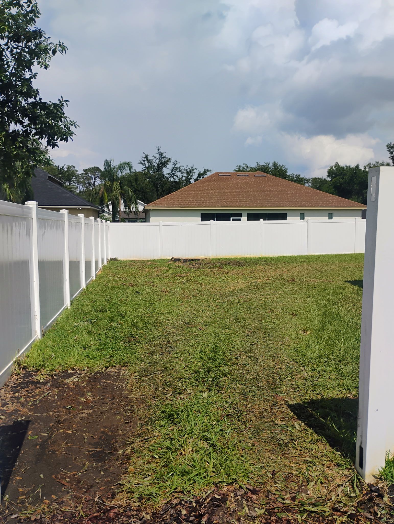 A backyard enclosed by white fences and a house with a brown roof in the distance. Cloudy sky.