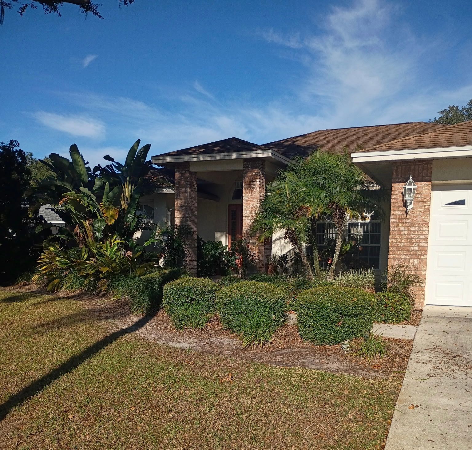 House with brick facade and covered porch, surrounded by green shrubs and trees, on a sunny day.