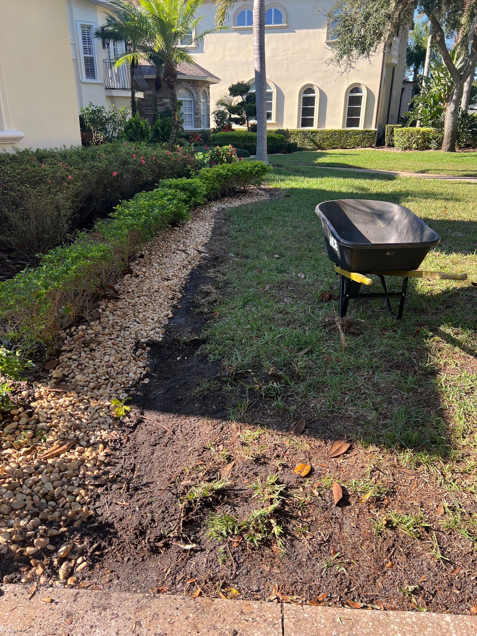 Wheelbarrow on grass next to a freshly mulched garden bed edged by a trimmed hedge. Houses in background.