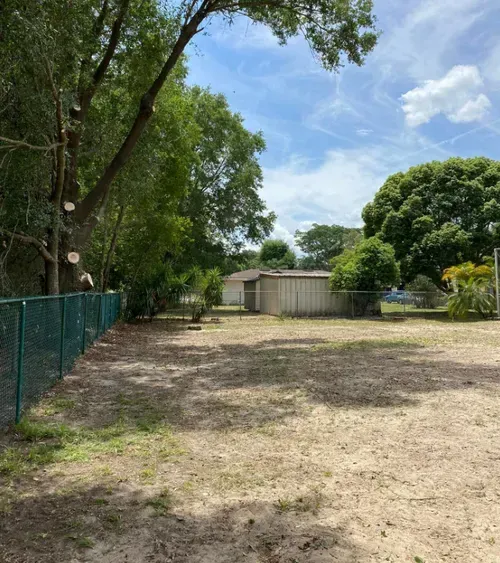 An empty dirt lot with a green fence and two metal sheds, under a bright, sunny sky.