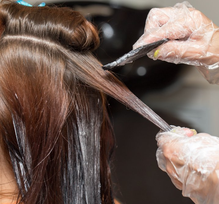 Hands in gloves applying hair dye with a brush to brown hair in a salon setting.