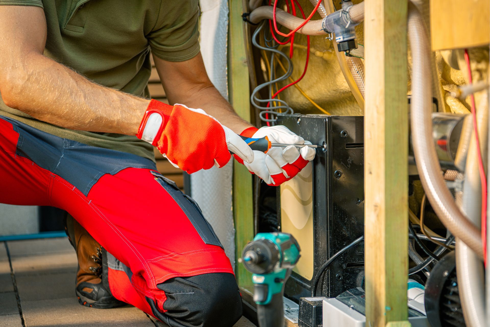 Person in red work pants and gloves using a screwdriver on machinery with wires.