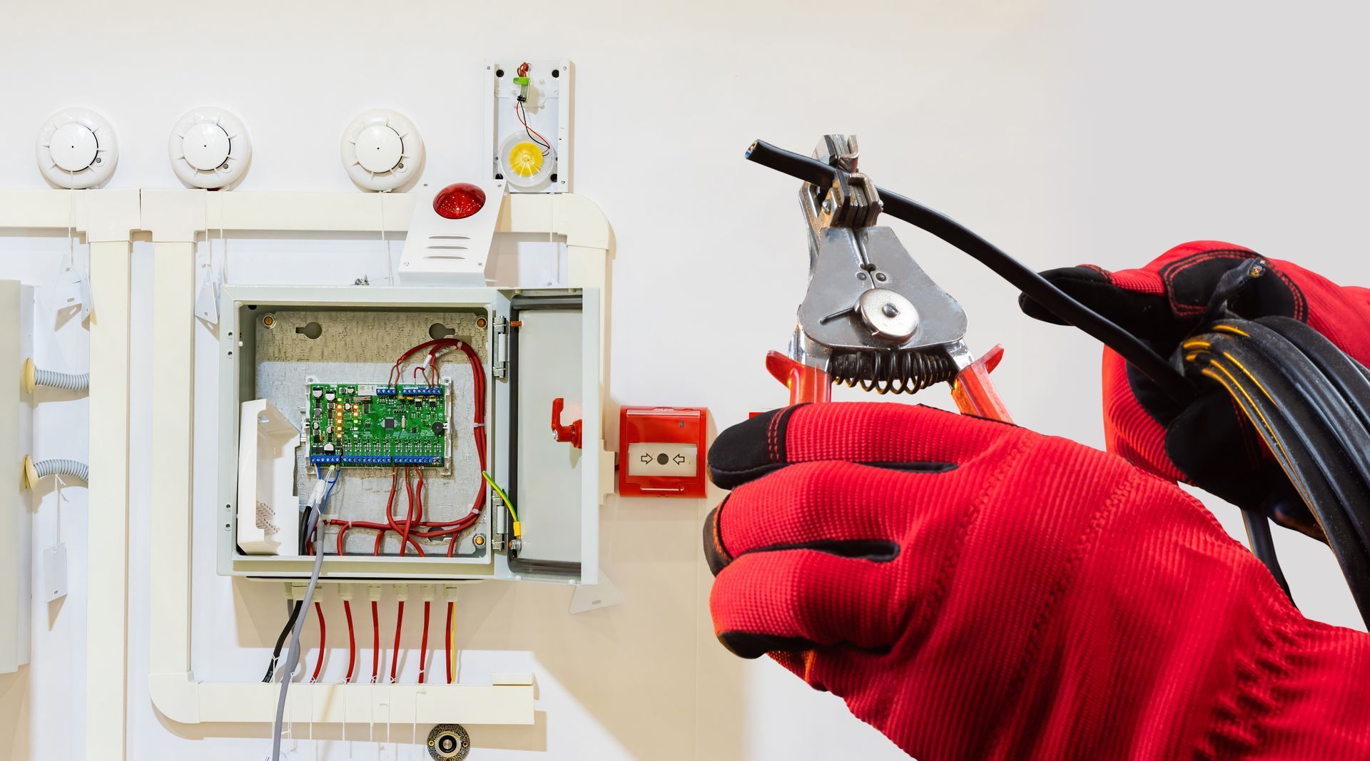 Electrician in red gloves using wire strippers on black cable near electrical panel.