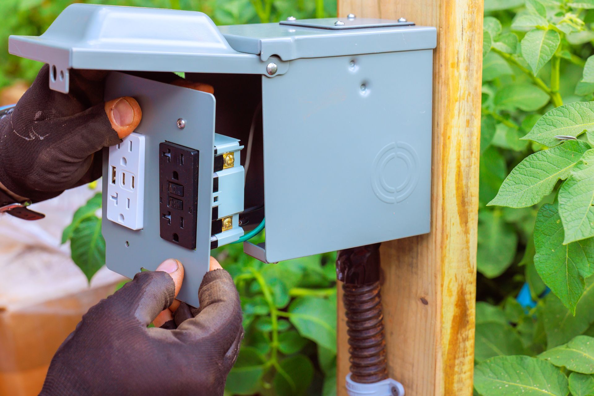 Hands installing electrical outlet in a gray weatherproof box mounted on a wooden post.