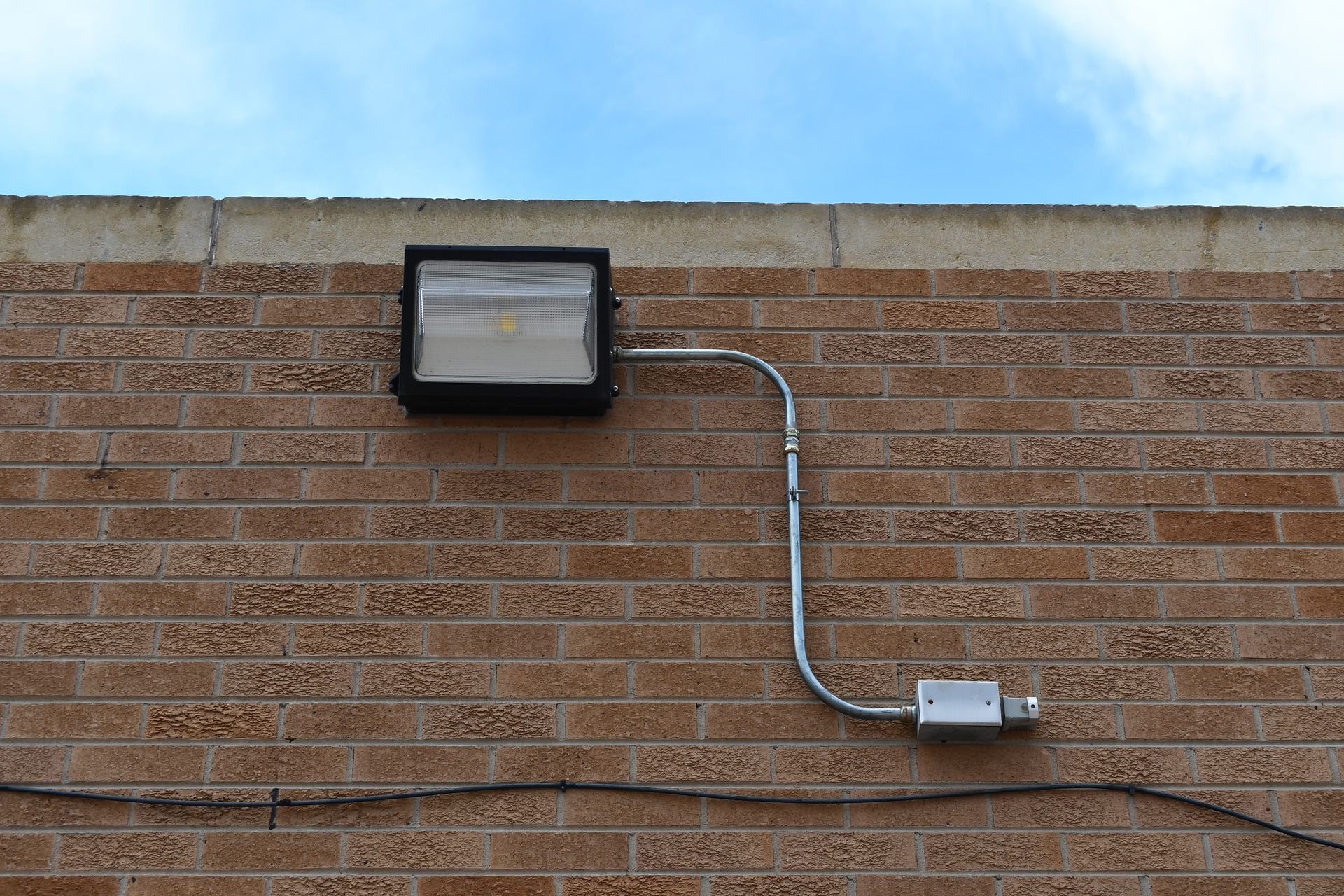 A black floodlight attached to a brick wall with conduit leading to a metal junction box.