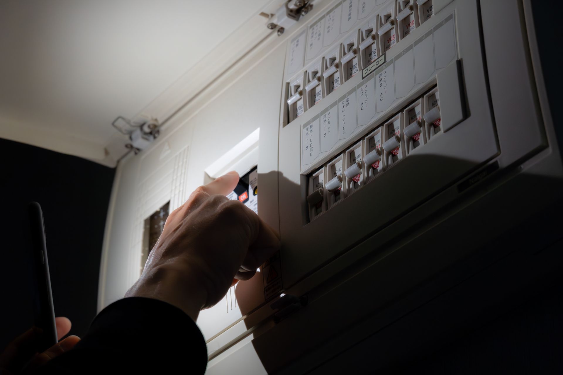 Person adjusting circuit breakers in a dimly lit electrical panel, using a phone as a flashlight.