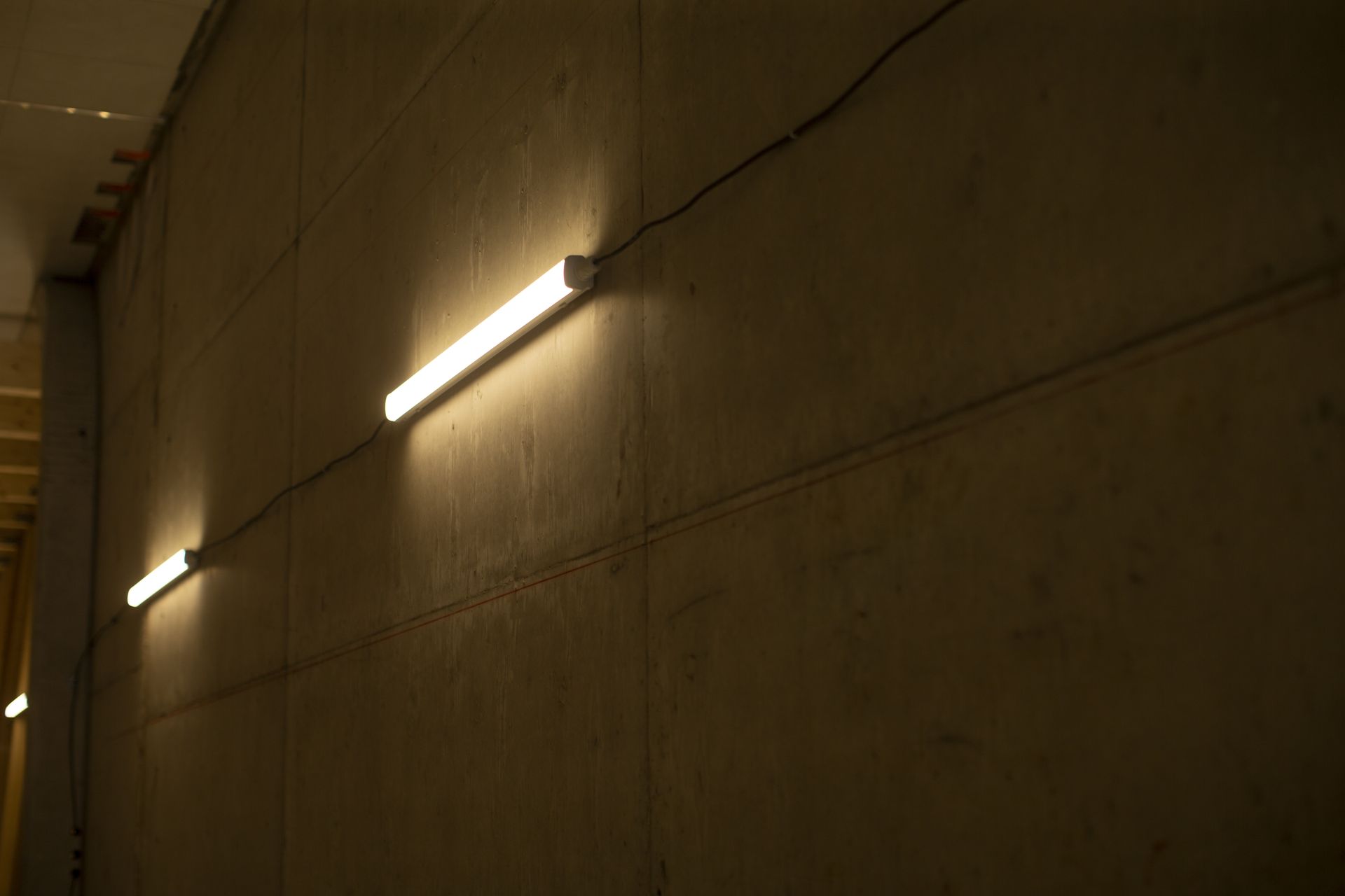 Two fluorescent lights illuminate a concrete wall in a dimly lit space.