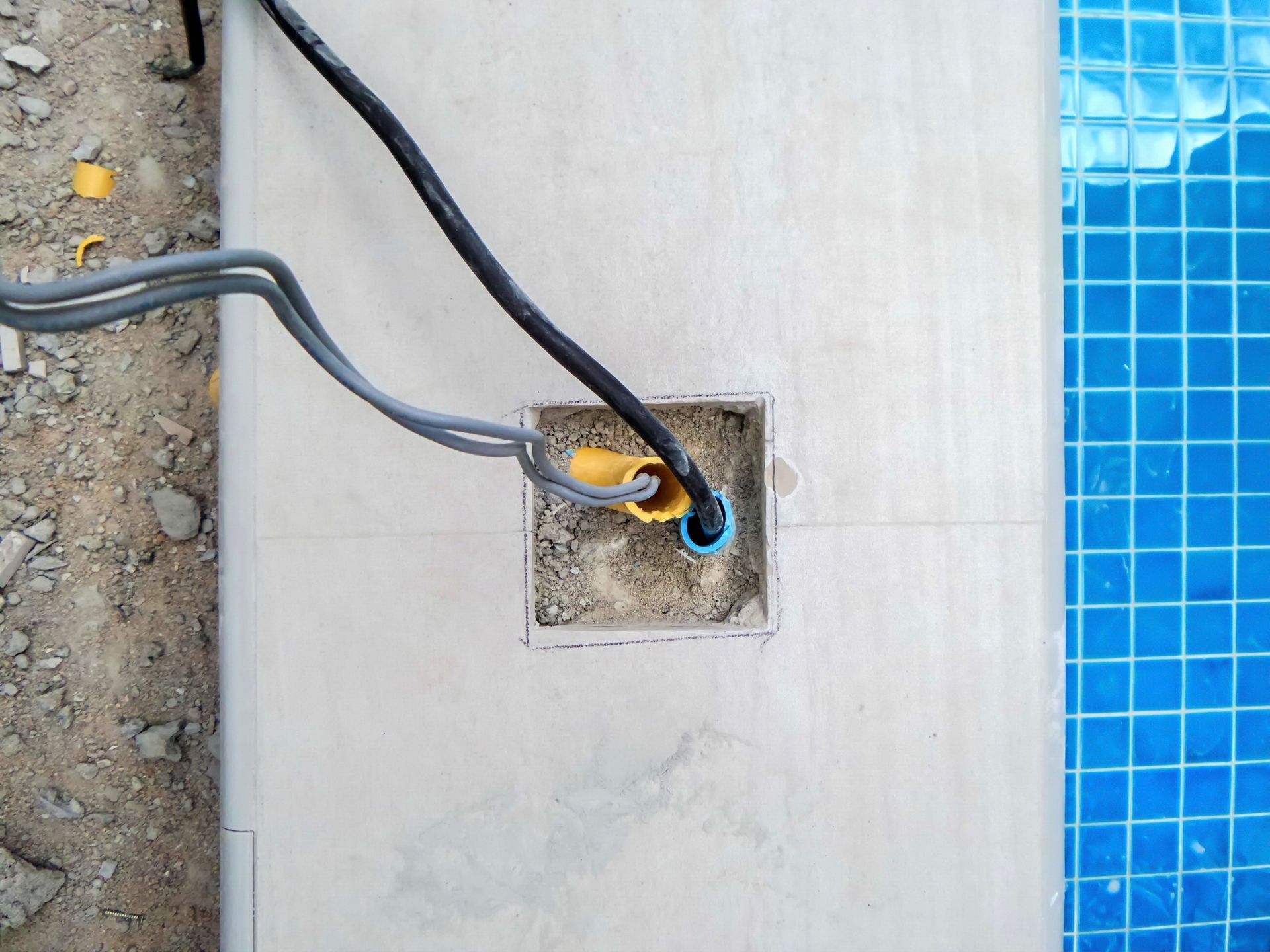 Electrical wires emerging from a square cut-out in a concrete surface next to a blue tiled pool.
