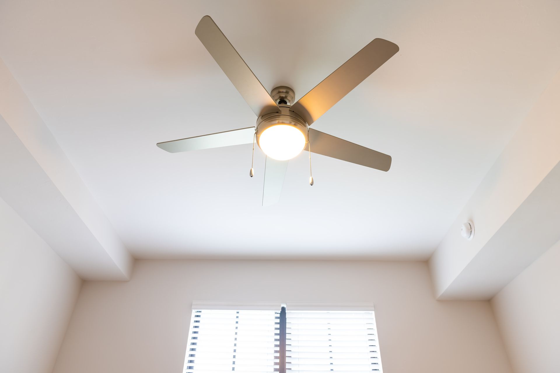Ceiling fan with light fixture centered on a white ceiling, above a window with blinds.