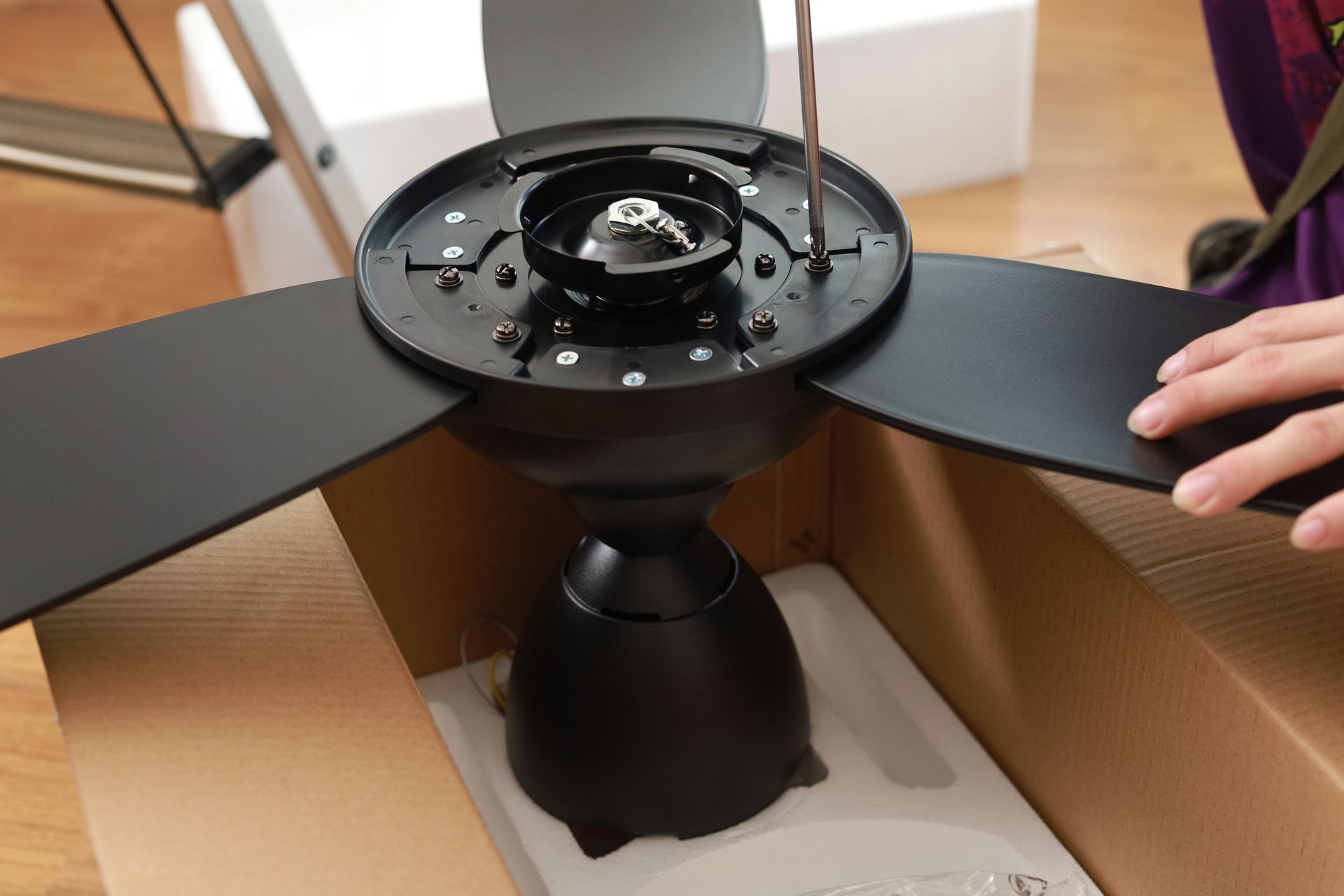 Black ceiling fan being assembled, inside a cardboard box. A hand is securing a blade with a screwdriver.