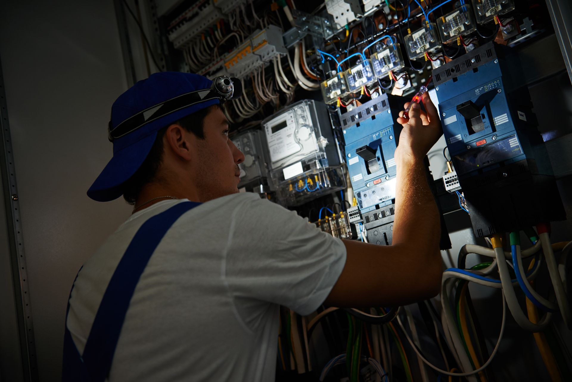 Electrician working on a circuit breaker panel, wearing a cap and headlamp.