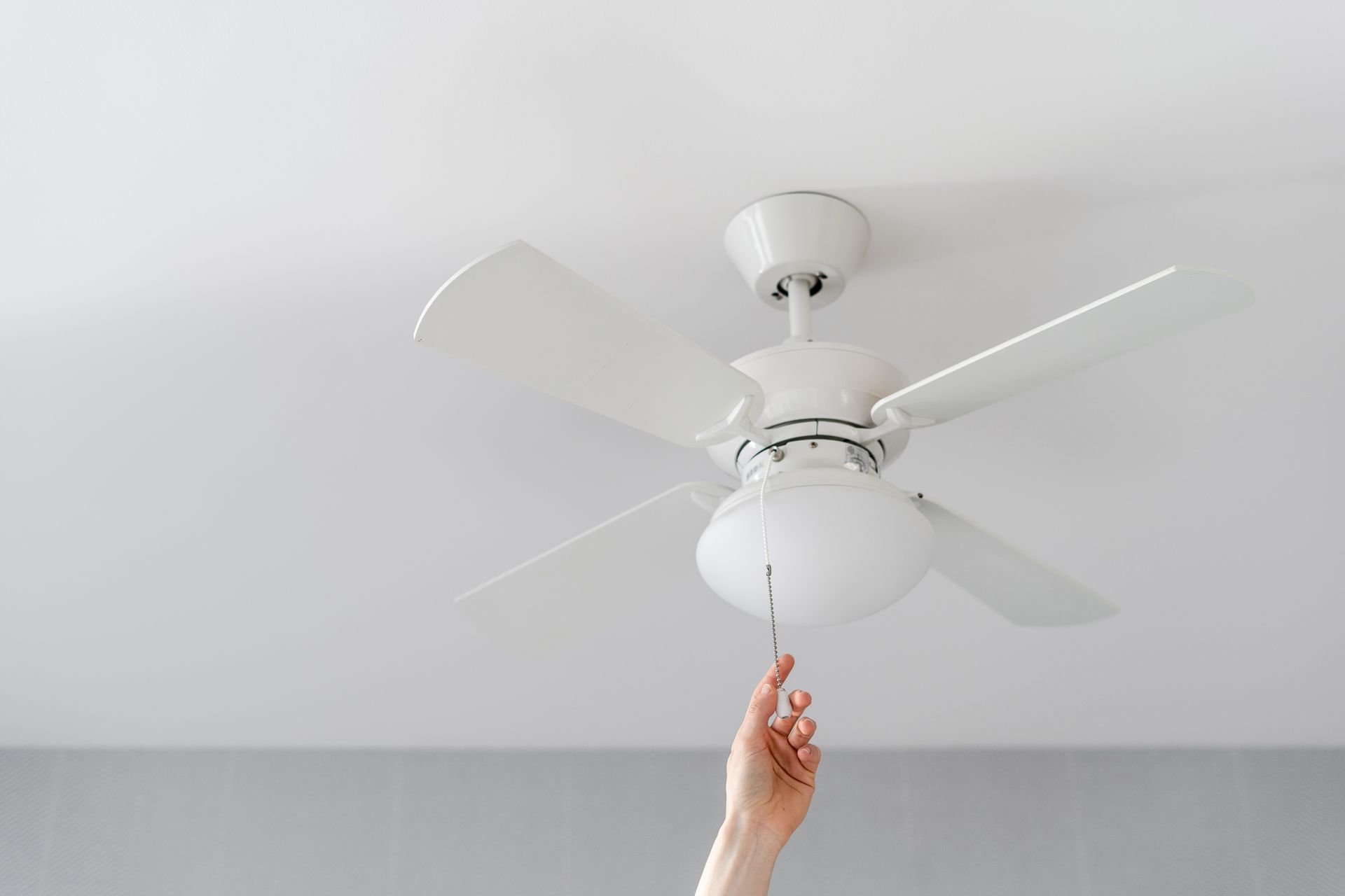 Hand pulling the cord of a white ceiling fan in a room with a white ceiling.