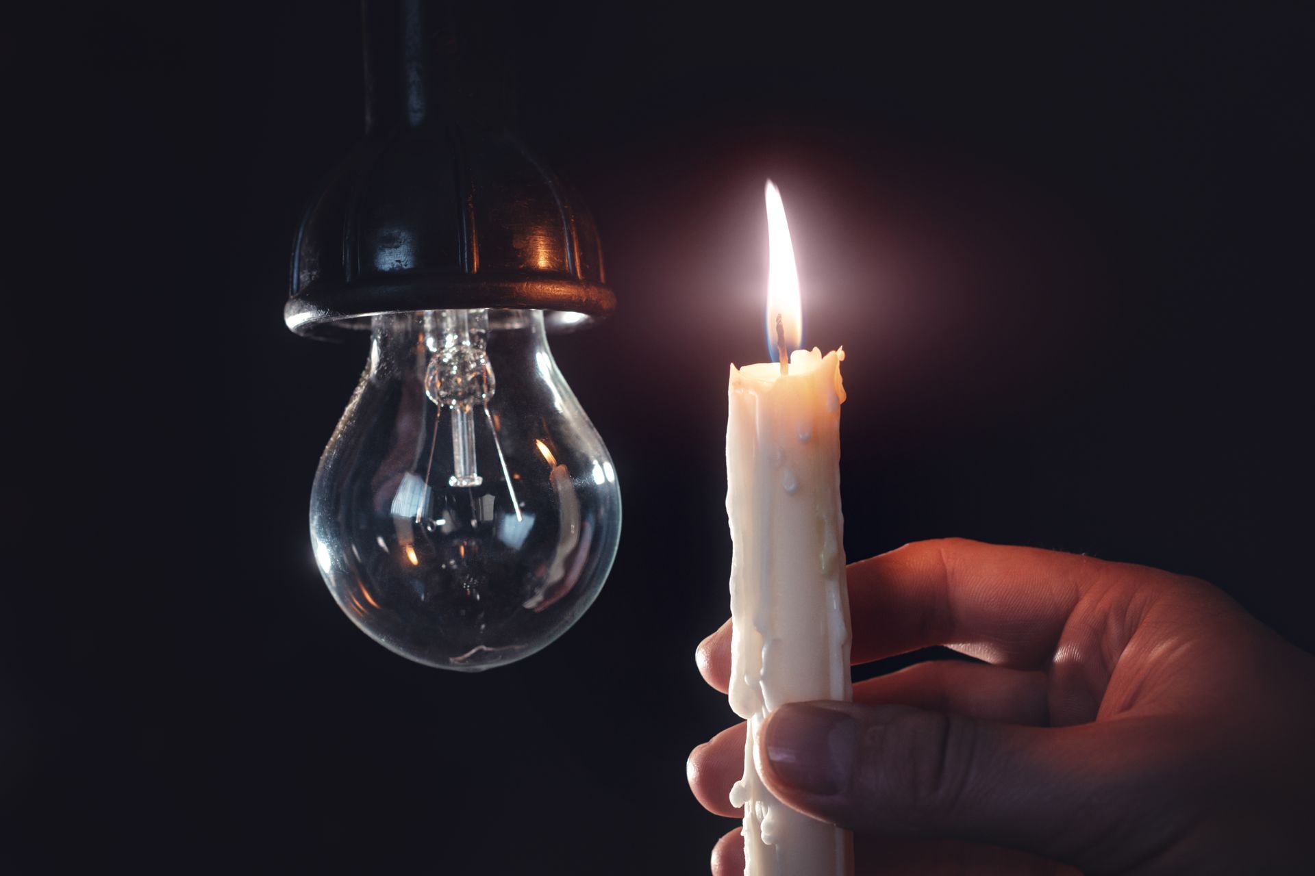 Hand holding a lit candle next to a non-glowing lightbulb in darkness.