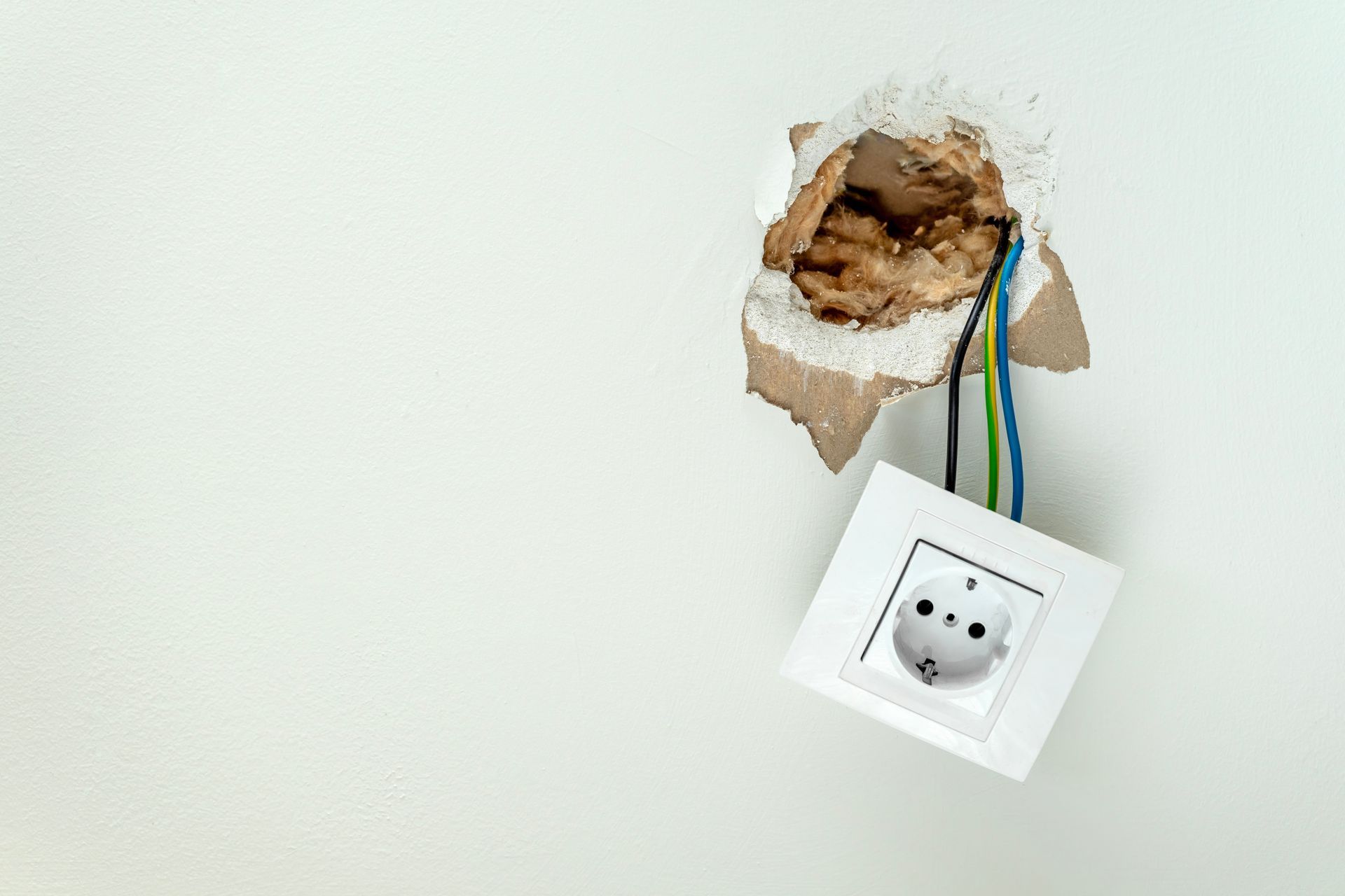 Damaged electrical outlet in a white wall; wires exposed.