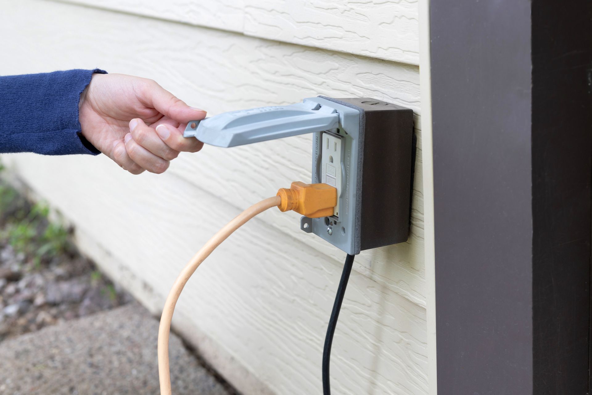 Hand opening a weatherproof outlet cover with an orange and beige cord plugged in. The outlet is mounted on siding.
